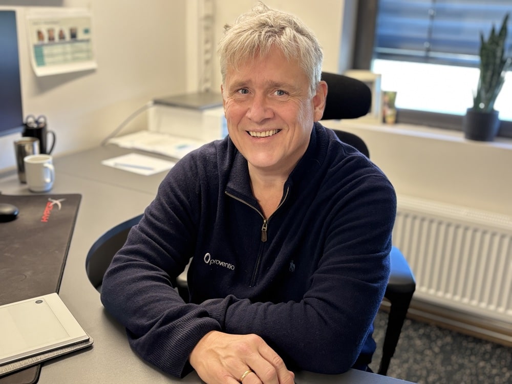 mann sitter i stol på kontor, armene på bordet. poserer, smiler.